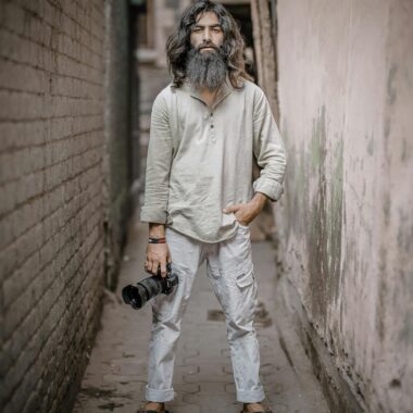 A bearded man holding a camera, standing confidently in a narrow alley.