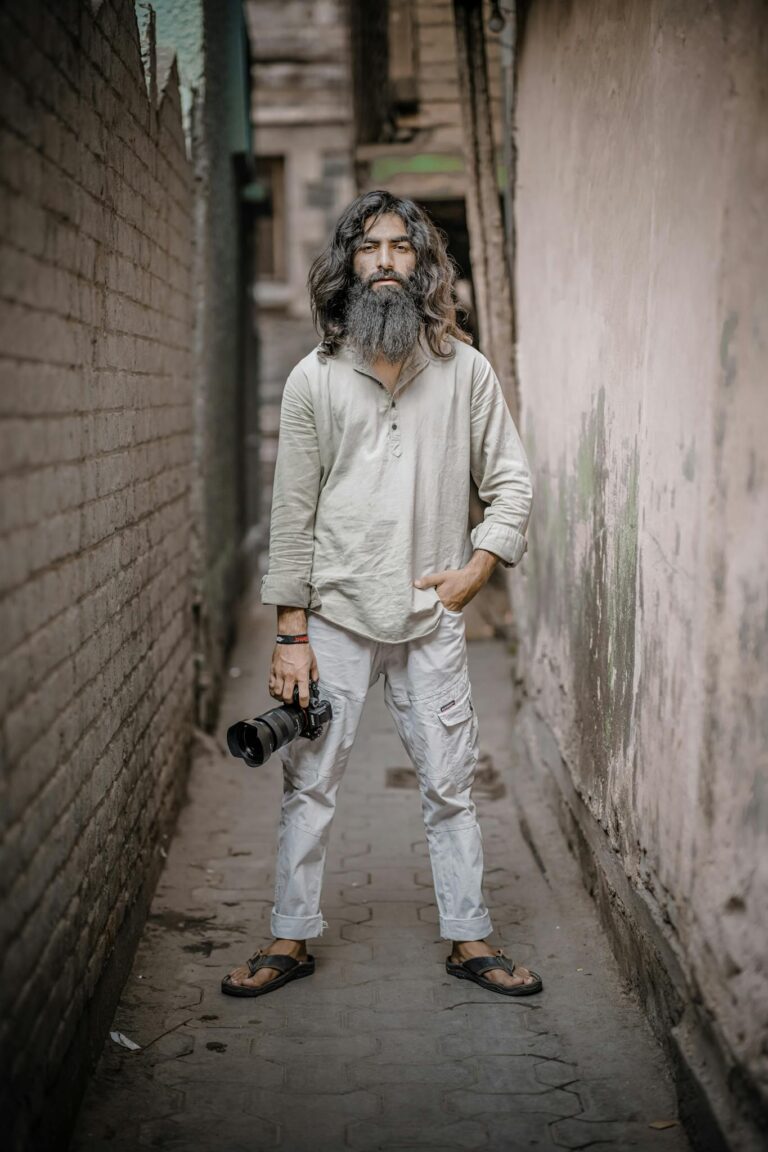 A bearded man holding a camera, standing confidently in a narrow alley.