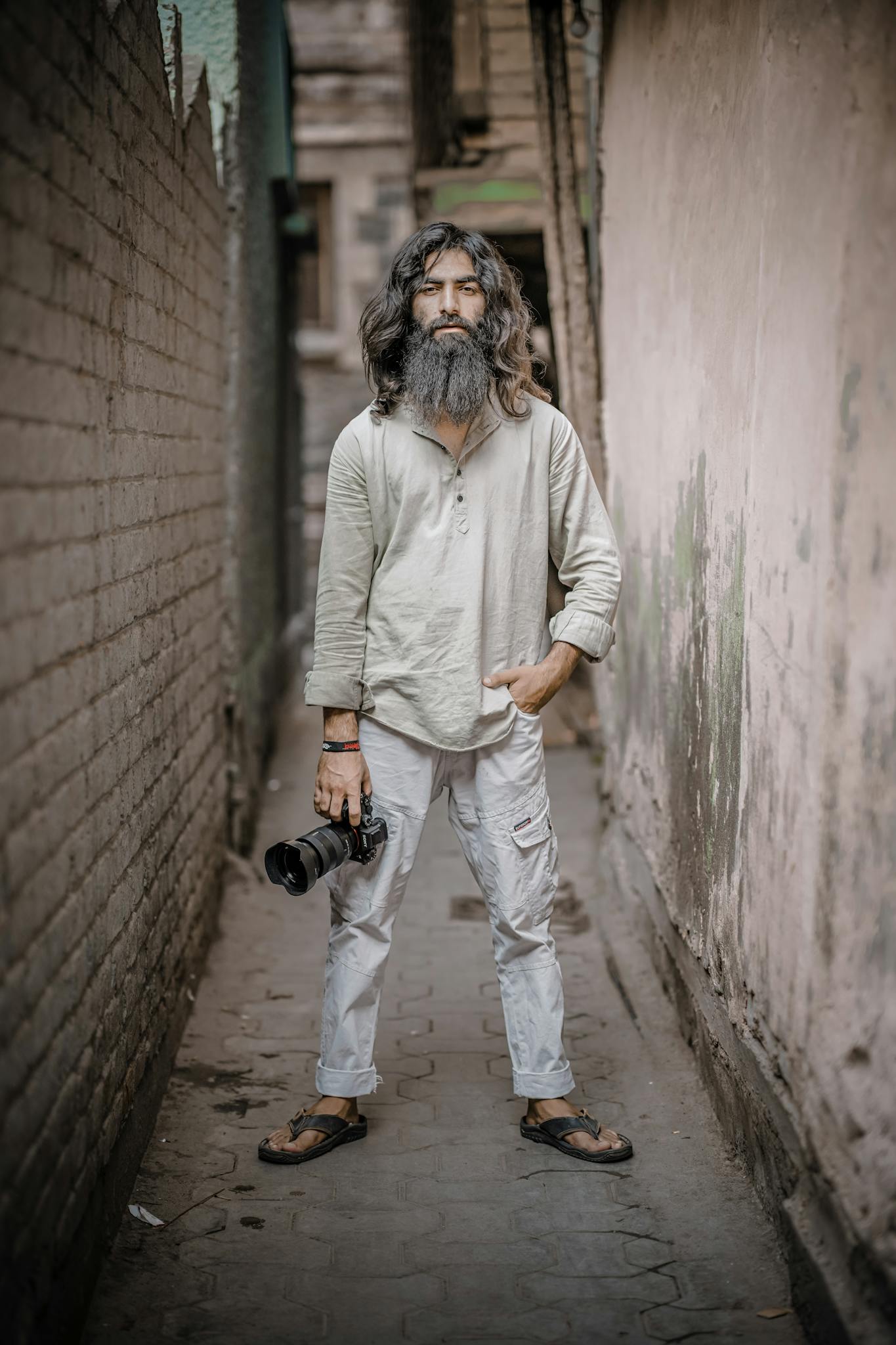 A bearded man holding a camera, standing confidently in a narrow alley.