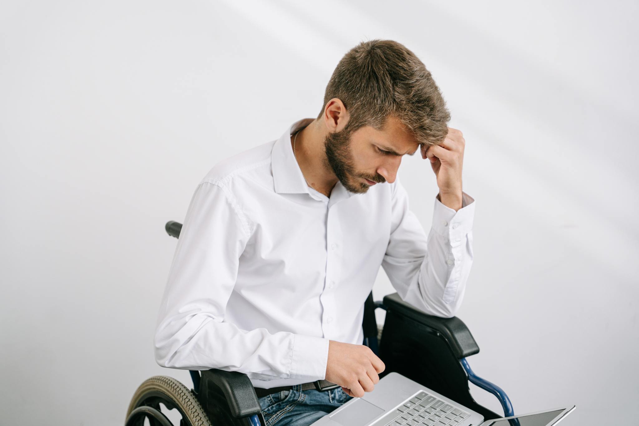 A young bearded man in a wheelchair works on his laptop indoors, wearing a white shirt.