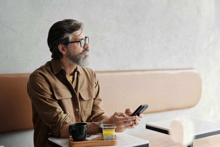 Casual bearded man enjoying coffee and using smartphone in a cozy caf&eacute;.