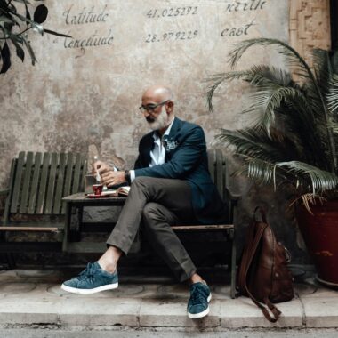 Full length of serious senior ethnic male with white beard in stylish outfit and eyeglasses drinking coffee while relaxing on wooden chair of authentic street cafe