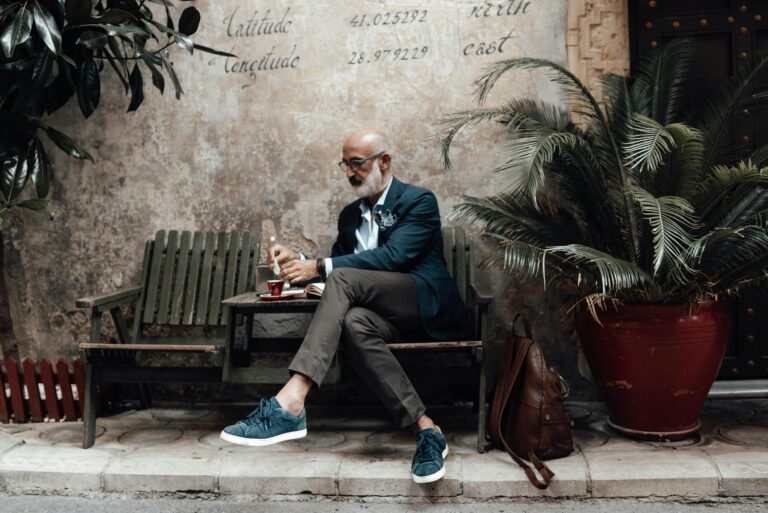 Full length of serious senior ethnic male with white beard in stylish outfit and eyeglasses drinking coffee while relaxing on wooden chair of authentic street cafe